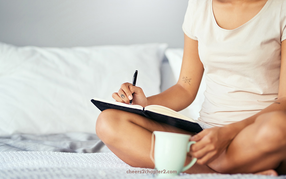 woman writing in a journal while in bed drinking coffee as part of her morning routine